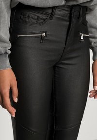 Svarta faux läderbyxor med fickor med dragkedja, en slät yta och en figurnära design med accentuerade sömmar längs sidorna.