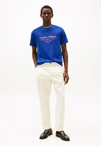 Blauwe katoenen t-shirt met witte en rode logo-print, gecombineerd met crèmekleurige broek. Model draagt zwarte instappers. Eenvoudige achtergrond.