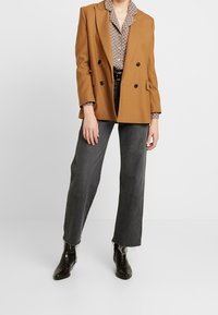Blazer marrone doppiopetto con rever a punta, camicia a fantasia sotto, abbinato a jeans grigi a gamba larga e stivaletti neri.
