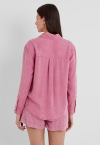 Pink denim shirt with long sleeves, buttoned cuffs, a back pleat, and a rounded hem, paired with matching pink shorts.