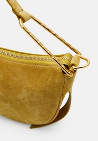 Mosterdgele suede schoudertas met een gladde textuur, voorzien van een gouden ritsafsluiting en een gedraaide handgreepbevestiging.