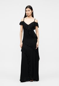 Νεαρή γυναίκα φοράει μαύρο μάξι φόρεμα με ακάλυπτους ώμους και καταρρακτώδεις πιέτες, μαζί με μαύρα σανδάλια, μπροστά σε απλό φόντο.