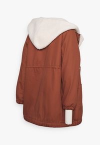 Chaqueta color óxido con capucha blanca y esponjosa. Cuenta con puños elásticos y un bolsillo lateral, diseñada con un ajuste relajado y una textura suave.