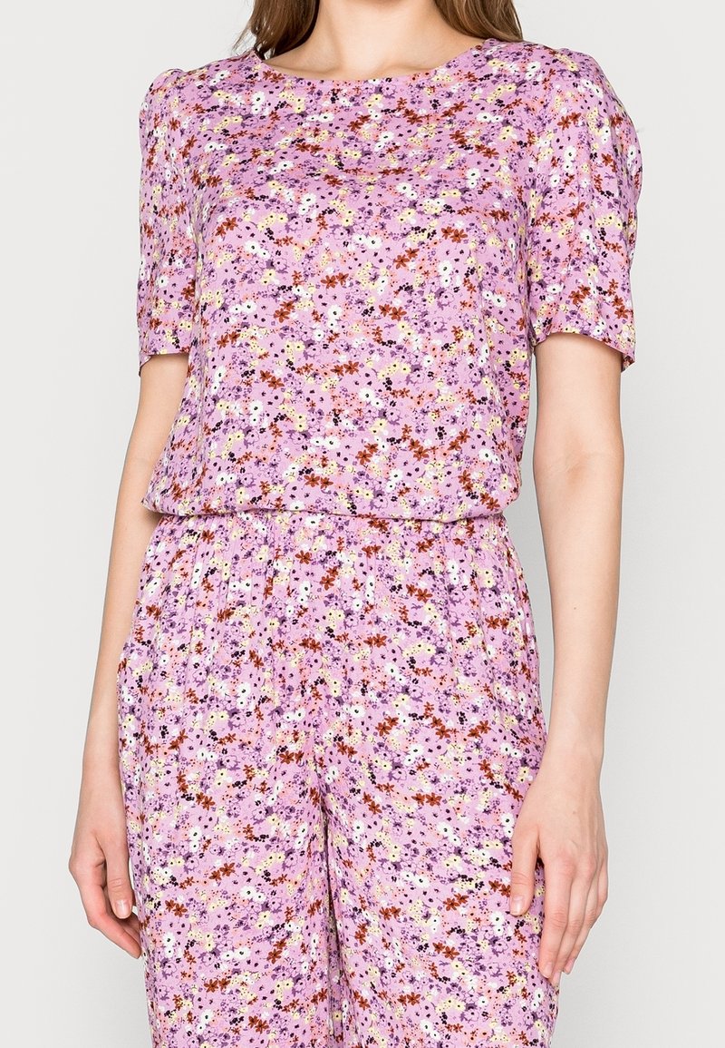 Femme portant un ensemble assorti top à manches courtes et pantalon avec un fond rose et un petit motif floral multicolore.