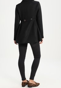 Cappotto nero su misura con design svasato, dettaglio a doppio bottone sul retro, abbinato a jeans skinny neri e ballerine con stampa leopardata.