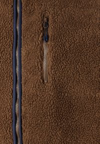 Chaqueta de forro polar marrón con una superficie texturizada, acentos en azul marino y un bolsillo con cremallera hecho de un tejido contrastante junto a la cremallera.