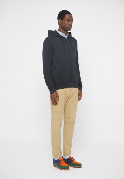 Σκούρα μπλε φούτερ με φερμουάρ και καγκουρό τσέπες, μπεζ τζιν και πολύχρωπα σουέτ παπούτσια με πορτοκαλί, μπλε και πράσινες λεπτομέρειες.