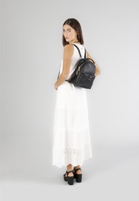 Svart läderryggsäck med guldfärgade detaljer; rund form, dragkedjestängning, framficka; bärs med en vit ögondroppsklänning och kraftiga svarta sandaler.