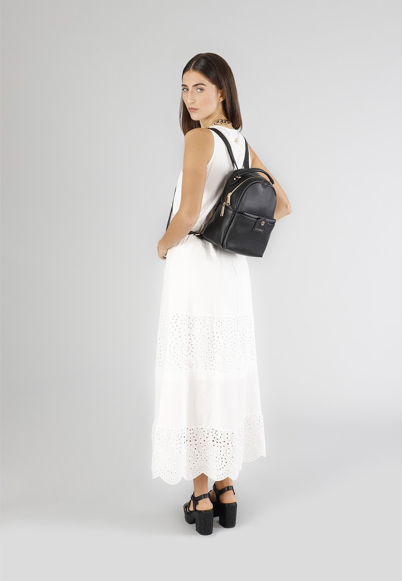 Svart läderryggsäck med guldfärgade detaljer; rund form, dragkedjestängning, framficka; bärs med en vit ögondroppsklänning och kraftiga svarta sandaler.