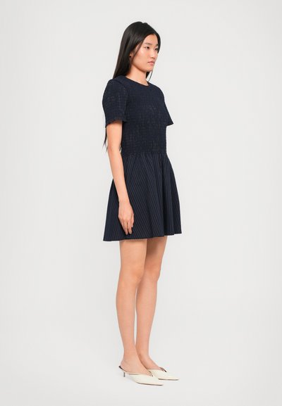 Sötétkék ruha texturált felső résszel és pliszírozott szoknyával. Rövid ujjak, kerek nyakkivágás és függőleges csíkmintázat a szoknyán.