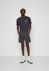 Camiseta de algodón gris con un parche de logo colorido, combinada con pantalones cortos grises. Zapatillas blancas con suelas negras y una gorra blanca completan el conjunto.