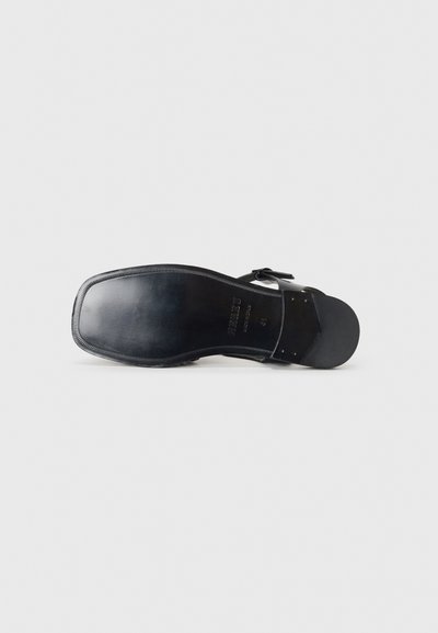 Suola di sandal in pelle nera con una texture liscia, forma arrotondata e un leggero tacco. Presenta un bordo cucito e dettagli in metallo.