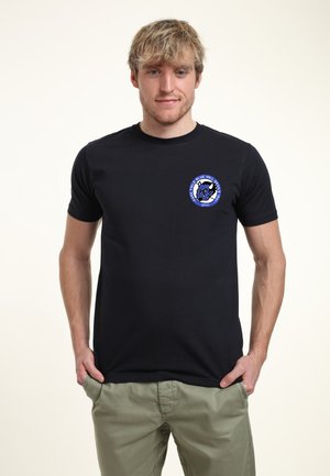 Černé tričko s krátkým rukávem s kulatým modrým grafickým logem na levé hrudi, zobrazujícím stylizovaný design zvířete.