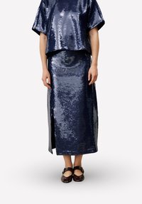 Marineblauw pailletten tweedelig outfit met een top in een losse pasvorm en een lange rok met een sleepscheur, gecombineerd met donkerbruine platte schoenen.