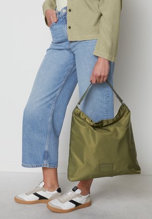 Olivgrüne Nylon-Tasche mit einer weichen, zerknitterten Textur, ausgestattet mit einer oberen Öffnung und einem breiten Tragegurt, gehalten von einer Person in hellblauen Jeans.