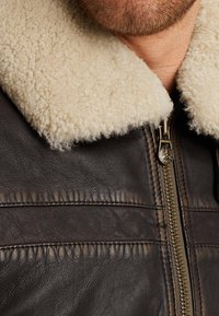 Brun læderjakke med kridhvid shearing-krave. Har en guld lynlås og synlig syning på kroppen. Det teksturerede stof viser tegn på slid.