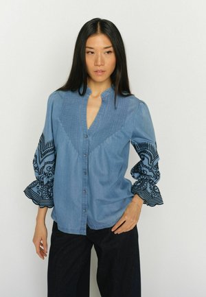 Kobieta w niebieskiej, zapinanej na guziki bluzce z czarnymi, wyszywanymi balonowymi rękawami i ciemnych, szerokich spodniach na tle jednolitego, białego tła.