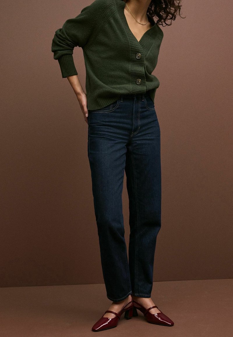 Femme portant un cardigan boutonné vert foncé, un jean bleu foncé et des chaussures à talons bordeaux, debout sur un fond marron.