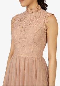 Femme portant une robe en dentelle sans manches rose poussiéreux avec un décolleté haut en forme de vagues et un motif floral sur le buste et la jupe en tulle.