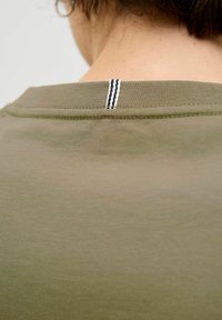 Alyvinės spalvos medvilnės marškinėliai su apvalia iškirpte, kuriuose yra plona juodos ir baltos siūlės detalė nugaros iškirptėje. Lygi tekstūra.