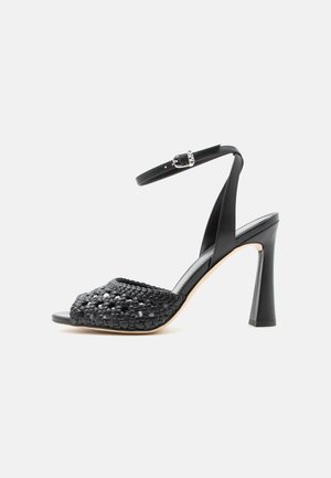 Sandalo nero con tacco alto, cinturino intrecciato sulla parte anteriore, cinturino alla caviglia con fibbia argentata e tacco largo svasato su sfondo neutro.