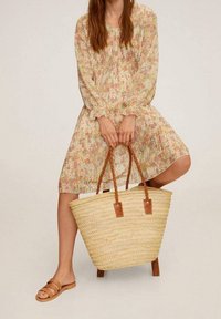 Strohtote mit gewebter Textur, braunen Lederakzenten und geflochtenen Griffen. Das Model trägt ein blumengemustertes Kleid und hellbraune Riemchensandalen.