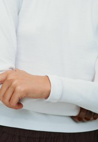 Bijela rebrasta majica dugih rukava, s uskim manžetama. Ruka je odložena na smeđi pojas s tri okrugla detalja.