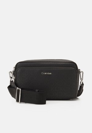 Black Calvin Klein crossbody bag with silver zippers, adjustable strap, and subtle embossed logo pattern on front pocket.