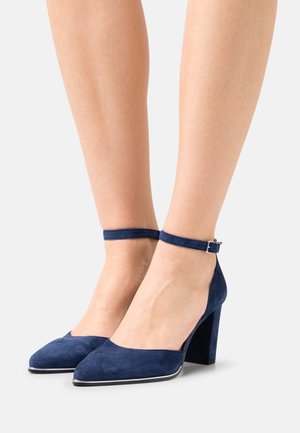 Gambe con indosso tacchi alti in suede blu navy con cinturini alla caviglia e fibbie argentate su uno sfondo bianco.