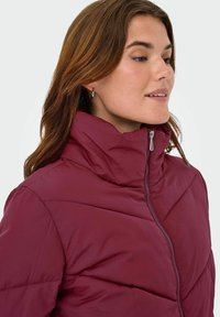 Bordo puf jakna s visokim ovratnikom, koja ima prednji zip i horizontalne šavne detalje za izolaciju i toplinu.