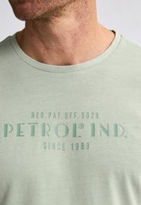 Camiseta de algodón verde claro con cuello redondo y un logo serigrafiado que dice "PETROL IND. SINCE 1989" en texto verde más oscuro.