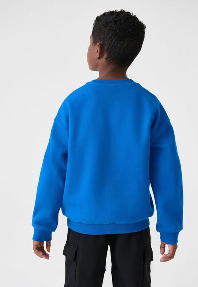 Modrá mikina s okrúhlym výstrihom a ribbovanými manžetami, s voľným strihom a jemnou textúrou, nosená cez čierne cargo nohavice.