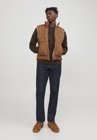 Brown quilted vest over a dark sweater, paired with dark denim jeans and brown suede shoes. Standing on a neutral background.