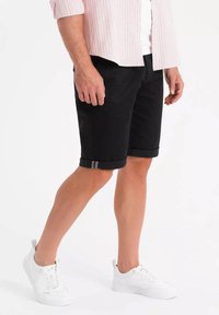 Shorts noirs avec des ourlets roulés, présentant un détail en fine rayure grise. Assortis avec des baskets blanches et une chemise à rayures claires à boutons.