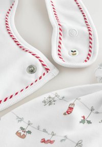Hvid bib med rød vævet kant, metaltrykknapper og et festligt mønster af julesokker, kristtorn og ornamenter trykt på stoffet.