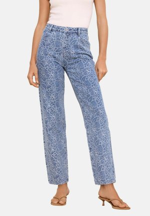 Kobieta nosząca niebieskie dżinsy z wysokim stanem z kwiatowym wzorem koronki, w połączeniu z beżowymi sandałami na obcasie i białym topem bez rękawów.