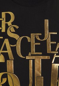 Black T-shirt featuring large, reflective gold letters with varying fonts and sizes, creating a layered text effect across the fabric.