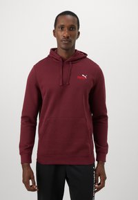 Homme portant un sweat à capuche maroon Puma avec une poche avant et un pantalon de survêtement noir Puma, se tenant contre un fond blanc uni.