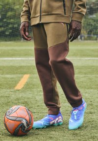 Un jogging marron et vert olive avec une texture douce, associé à des chaussures de football bleues et turquoises, à côté d'un ballon de football orange sur de l'herbe.