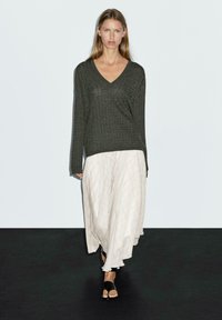 Dunkelgrüner, strukturiert gestrickter Pullover mit V-Ausschnitt, kombiniert mit einem hellen cremefarbenen, gemusterten Maxikleid. Getragen mit schwarzen Riemchensandalen.