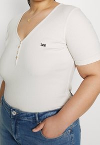 Bílý žebrovaný body s krátkým rukávem a výstřihem do V, vybavený funkčním zapínáním na knoflíky a černou výšivkou "Lee" na hrudi.