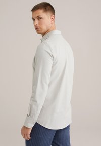 Camicia grigia chiaro a maniche lunghe con colletto, polsini abbottonati e una texture liscia. Indossata con pantaloni a righe navy.