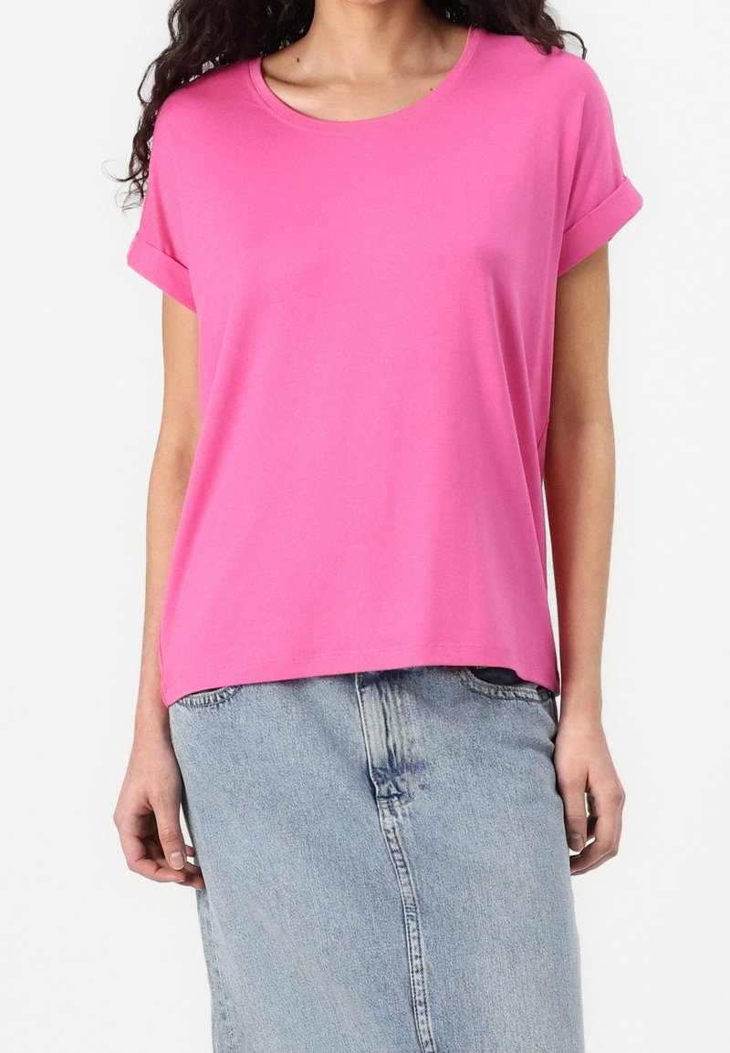 Femme portant un t-shirt rose vif à manches courtes avec manches retroussées et une jupe en jean bleue claire taille haute sur fond blanc.