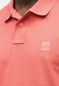 Πολο πουκάμισο σε κοραλλί ροζ με έναν κλειστό κουμπί και ανάγλυφο λογότυπο "BOSS" στην αριστερή περιοχή του στήθους.