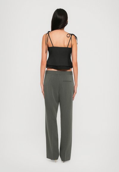 Woman with long dark hair wearing a black sleeveless top with tied straps and gray pinstripe wide-leg pants, standing facing away.