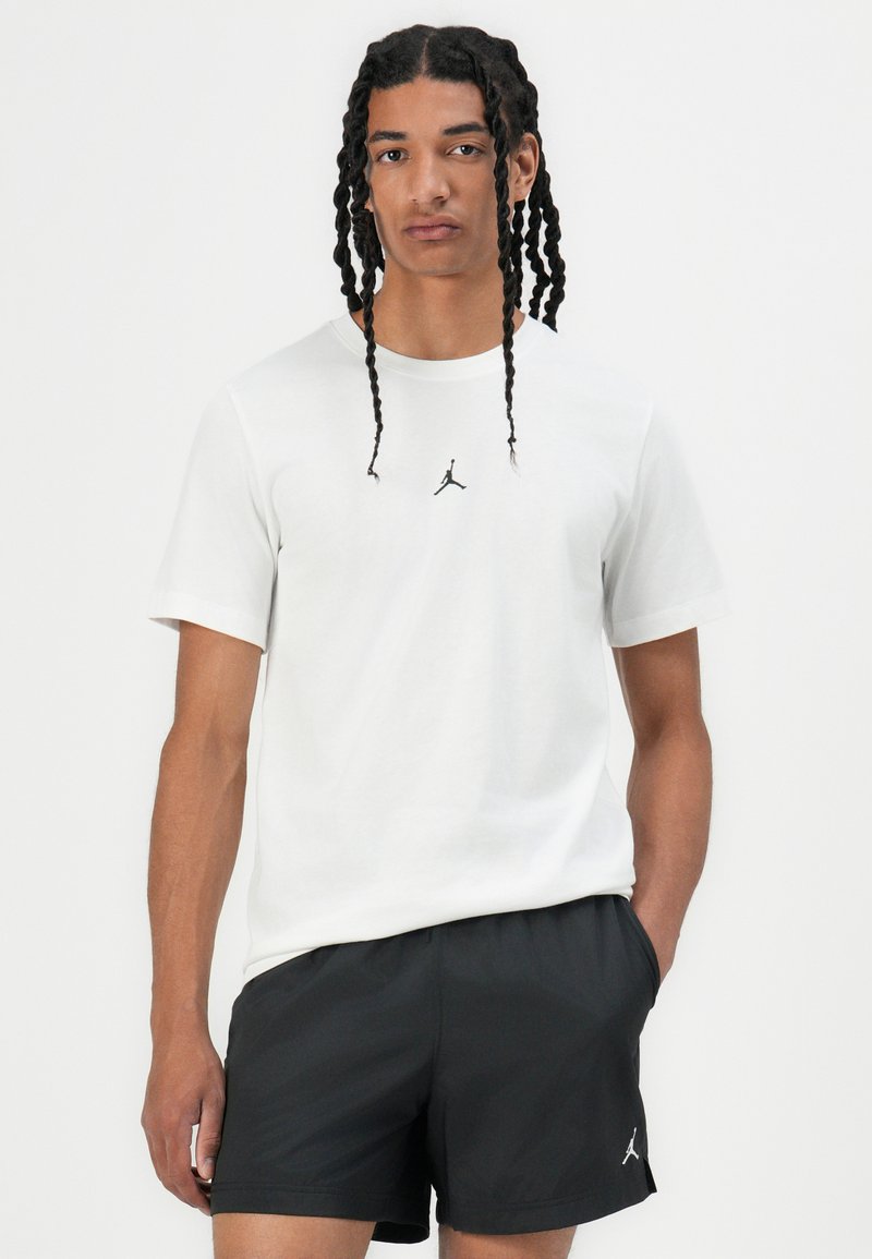 T-shirt en coton blanc avec un petit logo noir de Jordan au centre. Porté avec un short de sport noir ayant un logo sur la cuisse.