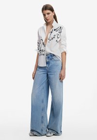Camicia bianca con stampe astratte nere, abbinata a jeans chiari a vita alta e gamba larga e sneakers bianche. Tessuto morbido, vestibilità casual.