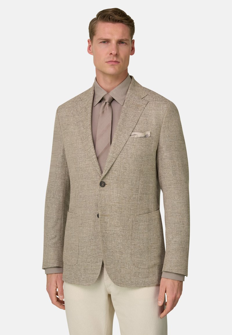 Homme portant un blazer beige texturé, une chemise marron clair, une cravate assortie, et un pantalon crème sur un fond uni.