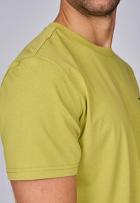 T-shirt à manches courtes en coton vert clair, avec un col rond et des détails de couture visibles le long des manches et de l'ourlet.