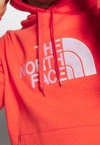 Felpa arancione brillante con logo bianco "The North Face" e lacci, indossata da una persona parzialmente visibile dal collo in giù.